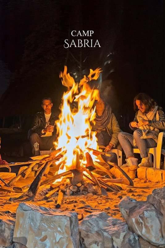 ضيوف يجلسون ليلًا حول نار مخيم مشتعلة على الرمل، وعبارة Camp Sabria في الأعلى