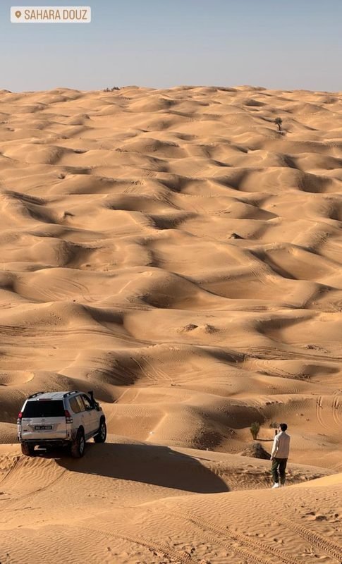 سيارة دفع رباعي بيضاء متوقفة على قمة كثيب فوق كثبان رملية لا نهاية لها، ورجل واقف بالقرب منها، وعبارة Sahara Douz في الأعلى