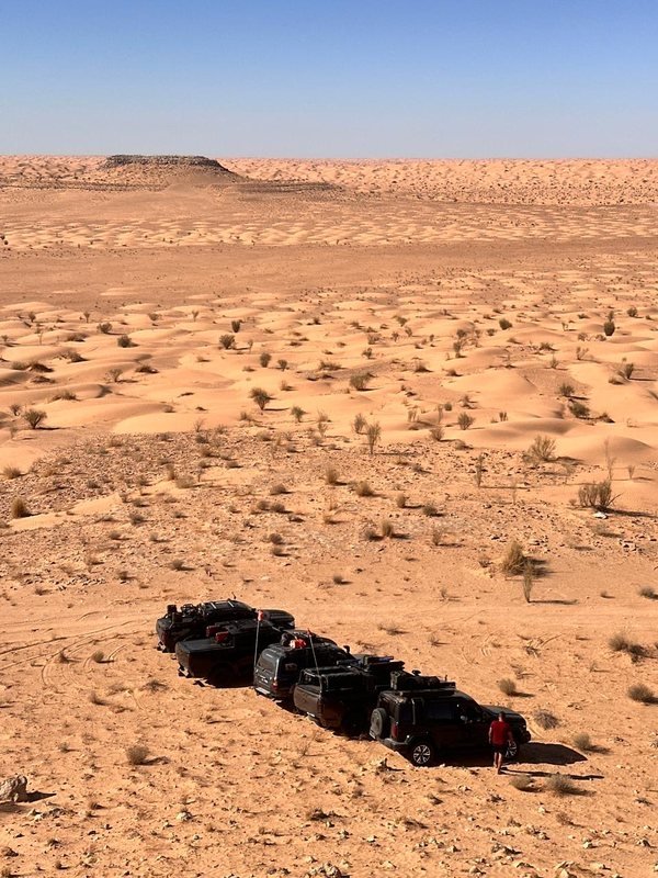 صف من سيارات الدفع الرباعي المتوقفة بين الكثبان الرملية، ورجل يمشي بالقرب منها، وهضبة مسطحة القمة في الأفق