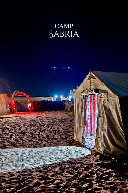 خيمة قماشية منفردة بستارة باب مخططة على الرمل ليلًا، خلفها قوس مضاء بالأحمر، وعبارة Camp Sabria في الأعلى